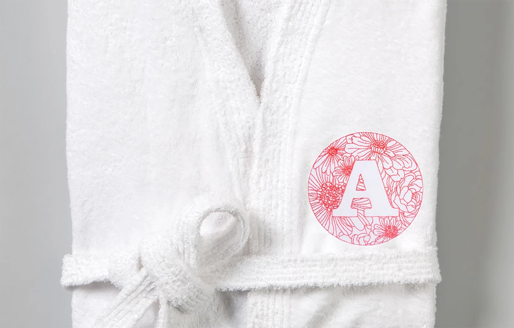 Folded white bath gown adorned with an intricately embroidered decorative letter 'A' monogram.